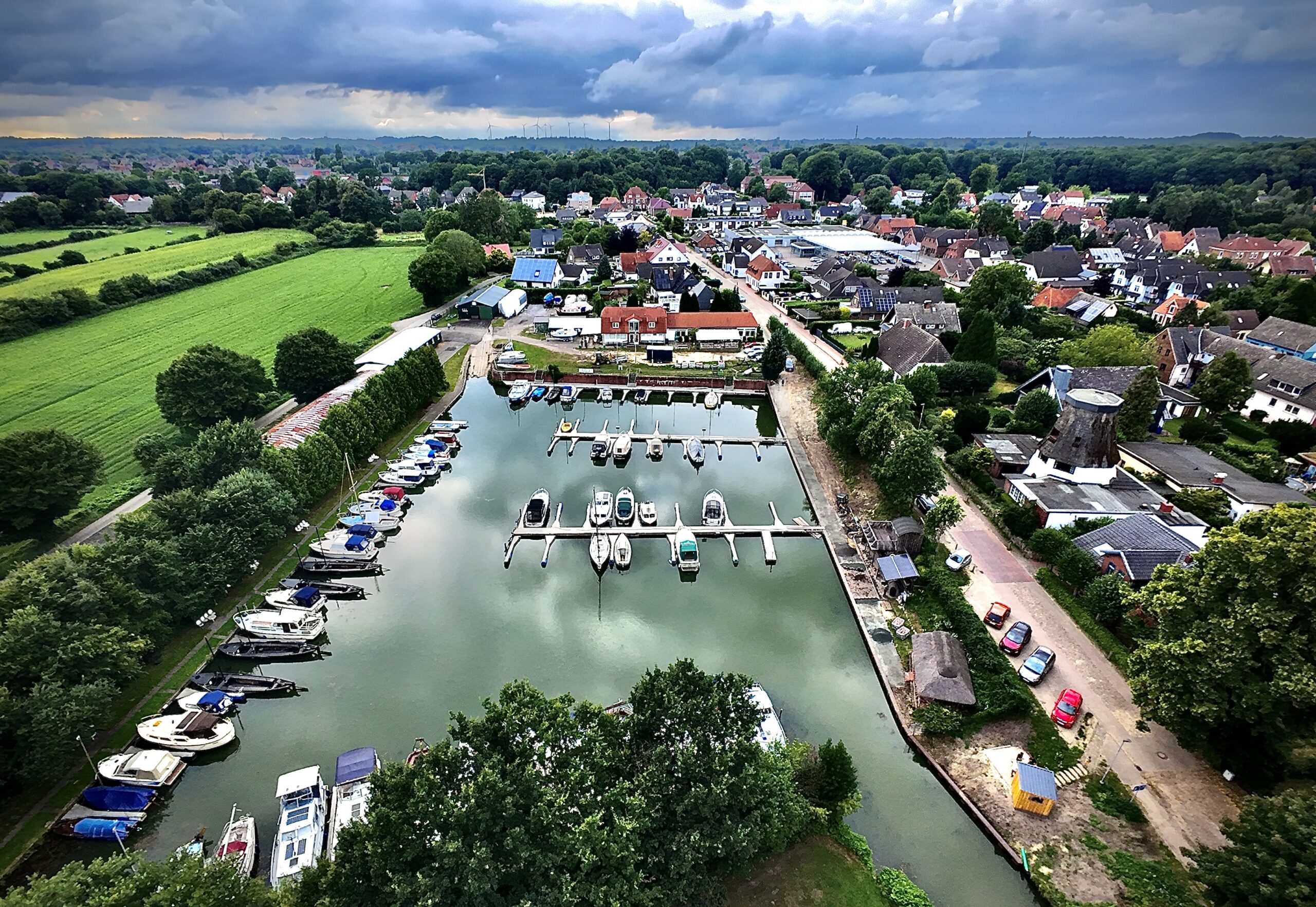 Das Foto ist mit einer Drohne aufgenommen worden. Es zeugt von oben einen kleinen Hafen in einer Stadt, im Hintergrund liegen Gebäude, Wälder und Wiesen