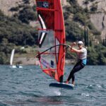 Ein Windsurfer mit Helm surft auf einem See vor hohen Felsen. Er reckt die Faust in die Luft und freut sich.