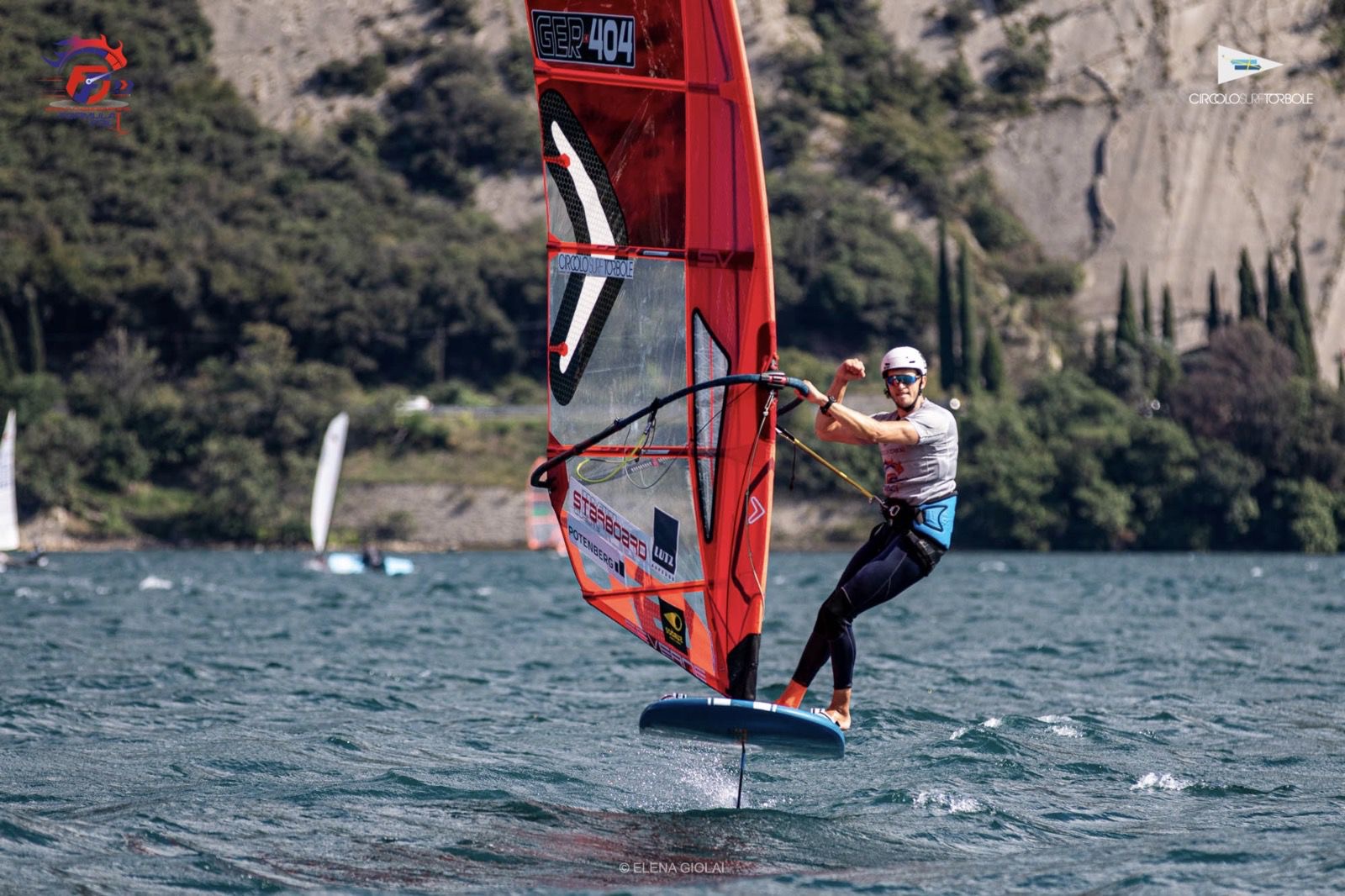 Ein Windsurfer mit Helm surft auf einem See vor hohen Felsen. Er reckt die Faust in die Luft und freut sich.