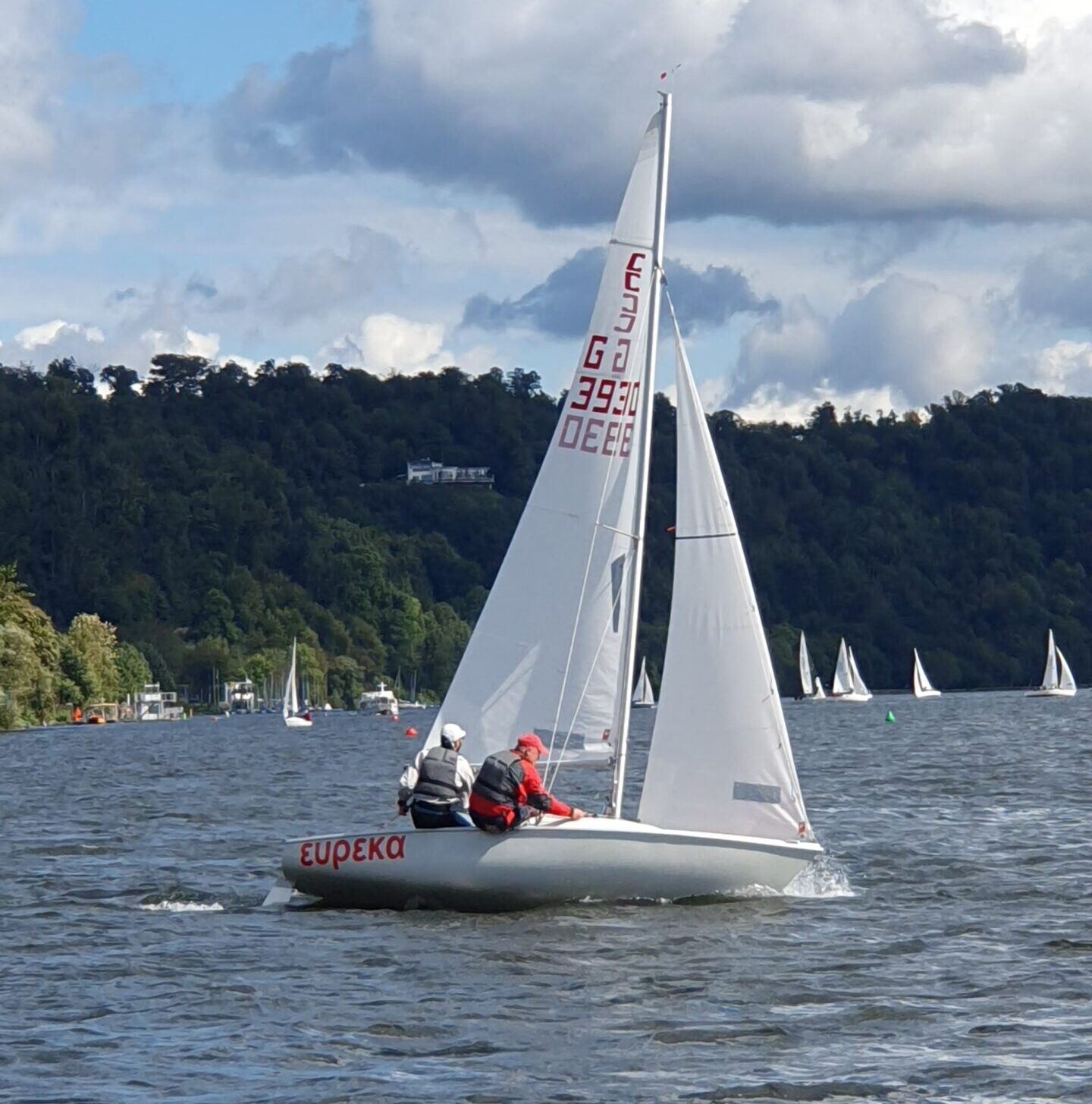 Eine Jolle der beliebten Conger-Klasse segelt auf einem See, im Hintergrund grüne Hügel, darüber Wolken und blauer Himmel.