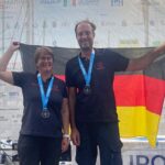 Eine Frau und ein Mann stehen auf einer Bühne mit einer Medaille um den Hals und halten eine Deutschlandflagge hoch.