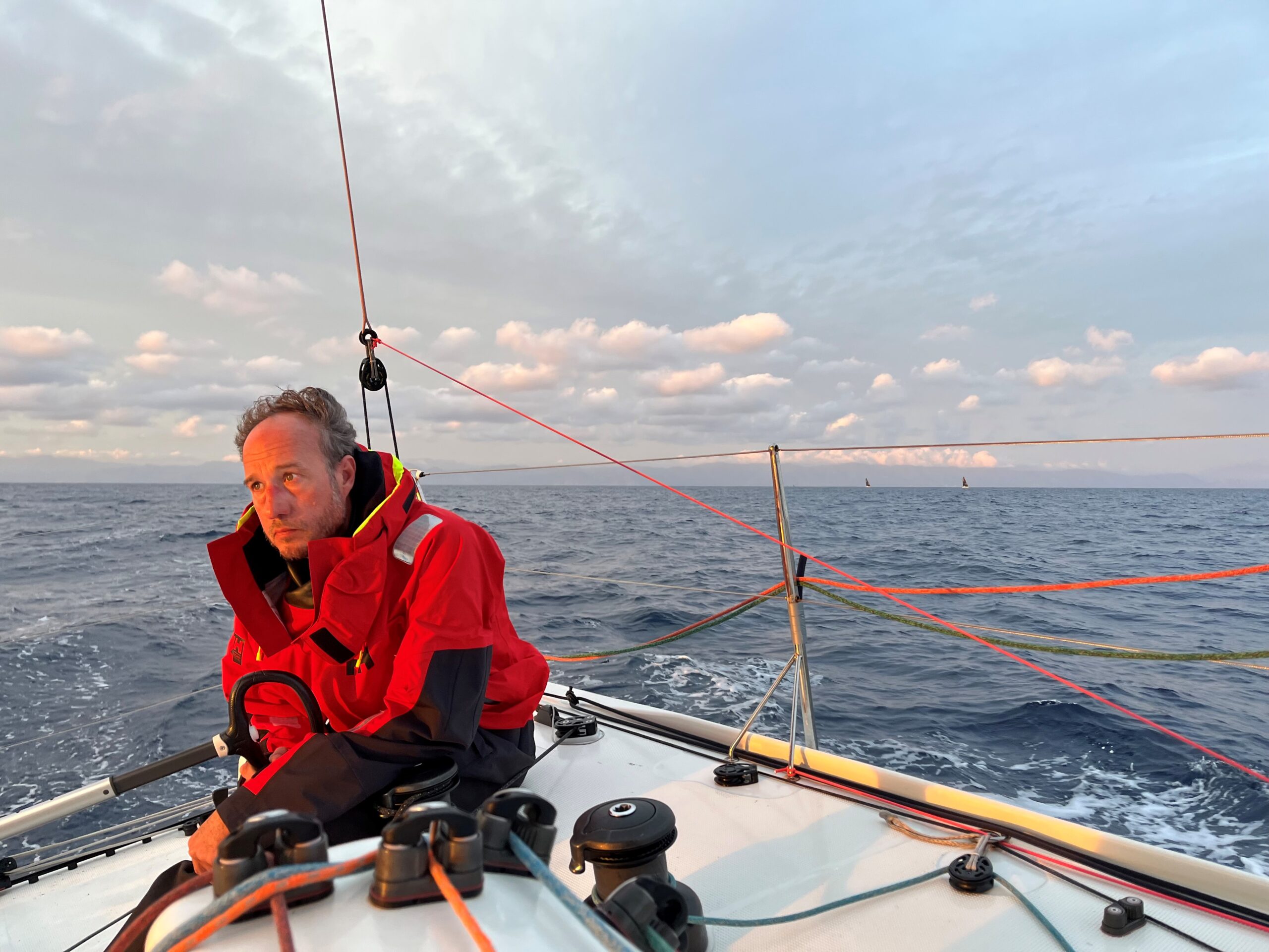 Ein Mann im Licht der untergehenden Sonne auf einem Segelboot, er trägt eine rote Jacke.