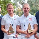 Zwei Männer mit Preisen von einer Regatta in der Hand, sie tragen weiße Polohemden und haben Medaillen um den Hals.