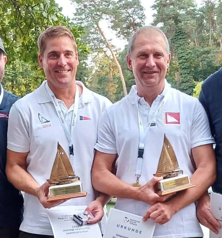 Zwei Männer mit Preisen von einer Regatta in der Hand, sie tragen weiße Polohemden und haben Medaillen um den Hals.