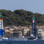 Zwei Katamarane mit den Flaggen von Deutschland und den USA vor Saint Tropez dahinter ein dunkler Pinienwald.
