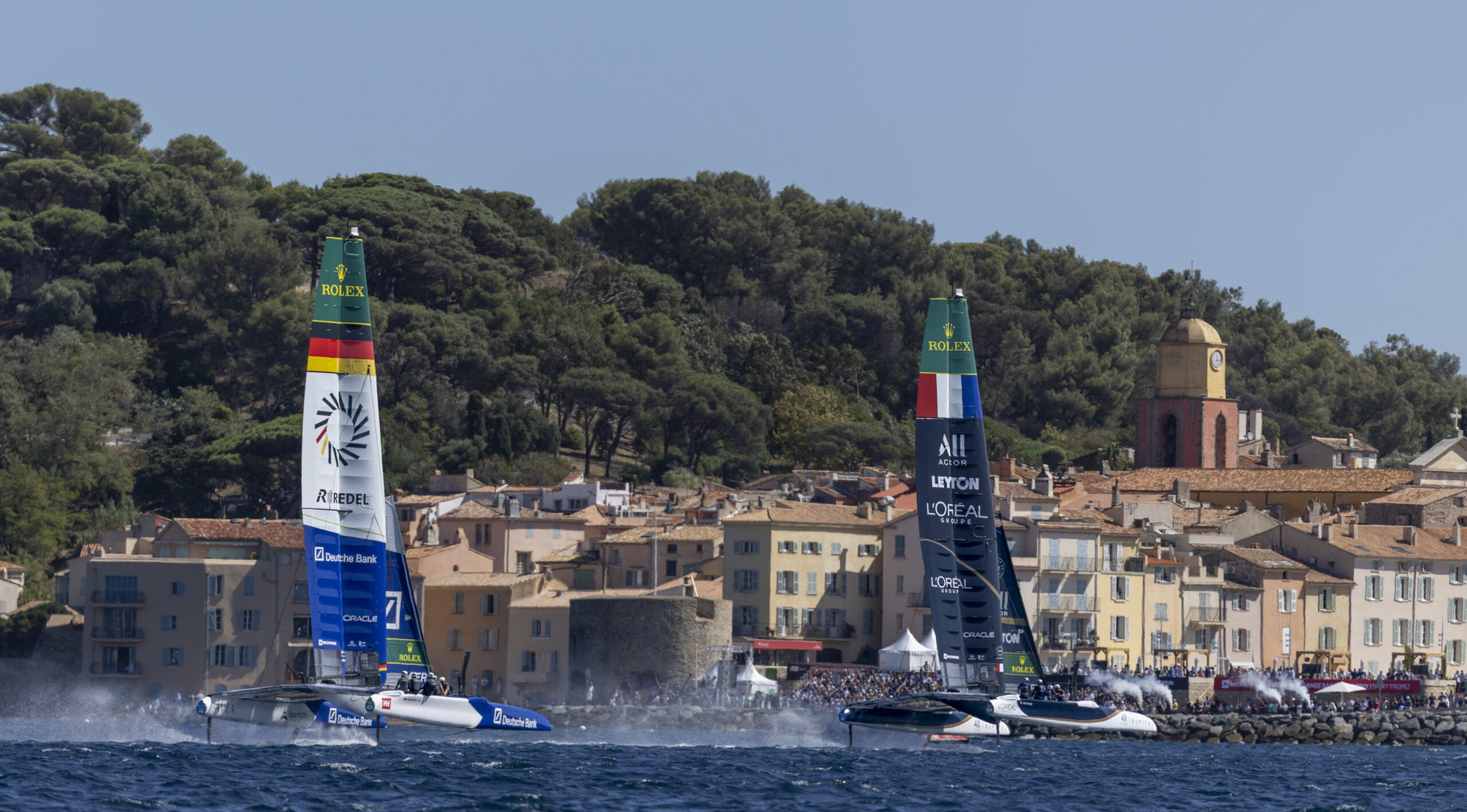 Zwei Katamarane mit den Flaggen von Deutschland und den USA vor Saint Tropez dahinter ein dunkler Pinienwald.