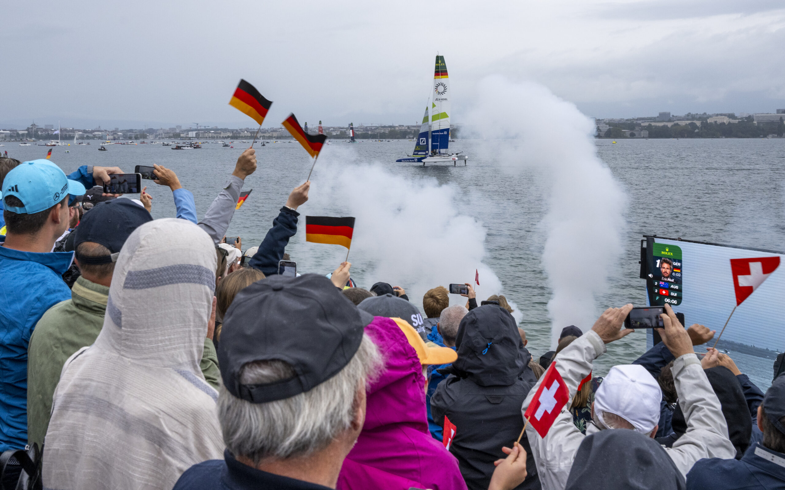 Eine Katamaran mit der deutschen Nationalflagge und dem Schriftzug der Deutschen Bank wird von Zuschauern mit deutschen und Schweizer Flaggen in den Händen bejubelt.