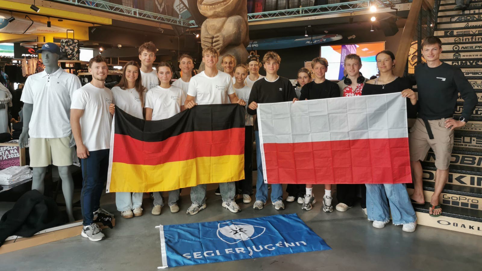 14 deutsche und polnische Jugendliche stehen in einem großen Surfshop. Sie lächeln in die Kamera und halten eine deutsche und eine polnische Flagge in der Hand.