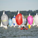 Schiffe mit bunten Spinnakern auf einem See davor eine gelbe Regattatonnen und ein rotes Schlauchboot.