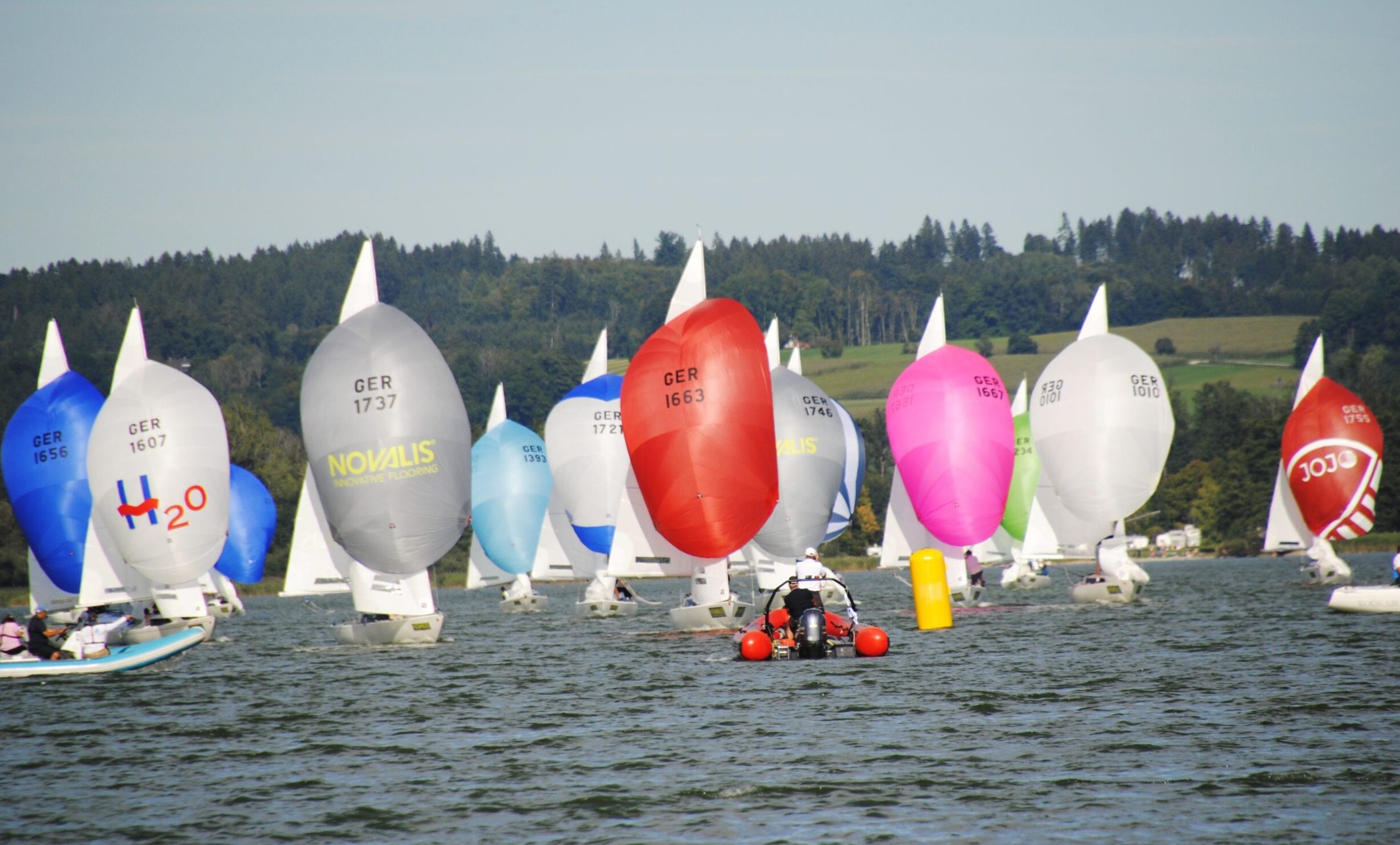 Schiffe mit bunten Spinnakern auf einem See davor eine gelbe Regattatonnen und ein rotes Schlauchboot.