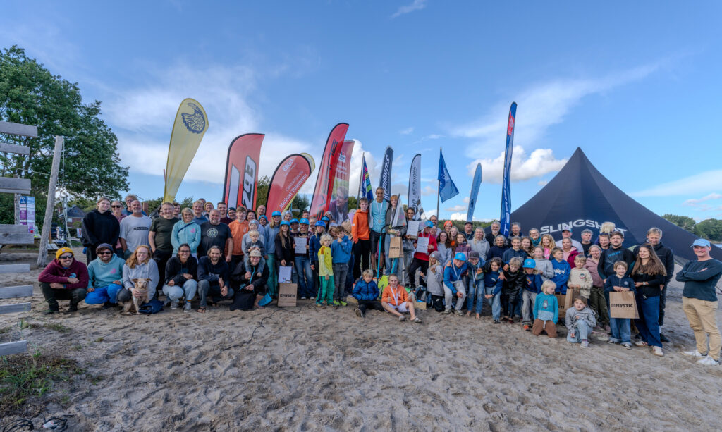 Viele junge Surferinnen und Surfer haben sich zu einem Gruppenfoto zusammengestellt. Sie sitzen und stehen am Strand.