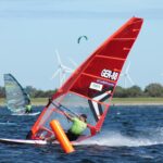 Ein Speedwindsurfer rast über die ostsee. Das Segel ist rot. Im Hintergrund ist ein zweiter Fahrer zu sehen, am Himmel sind Drachen von Kitesurfenden.