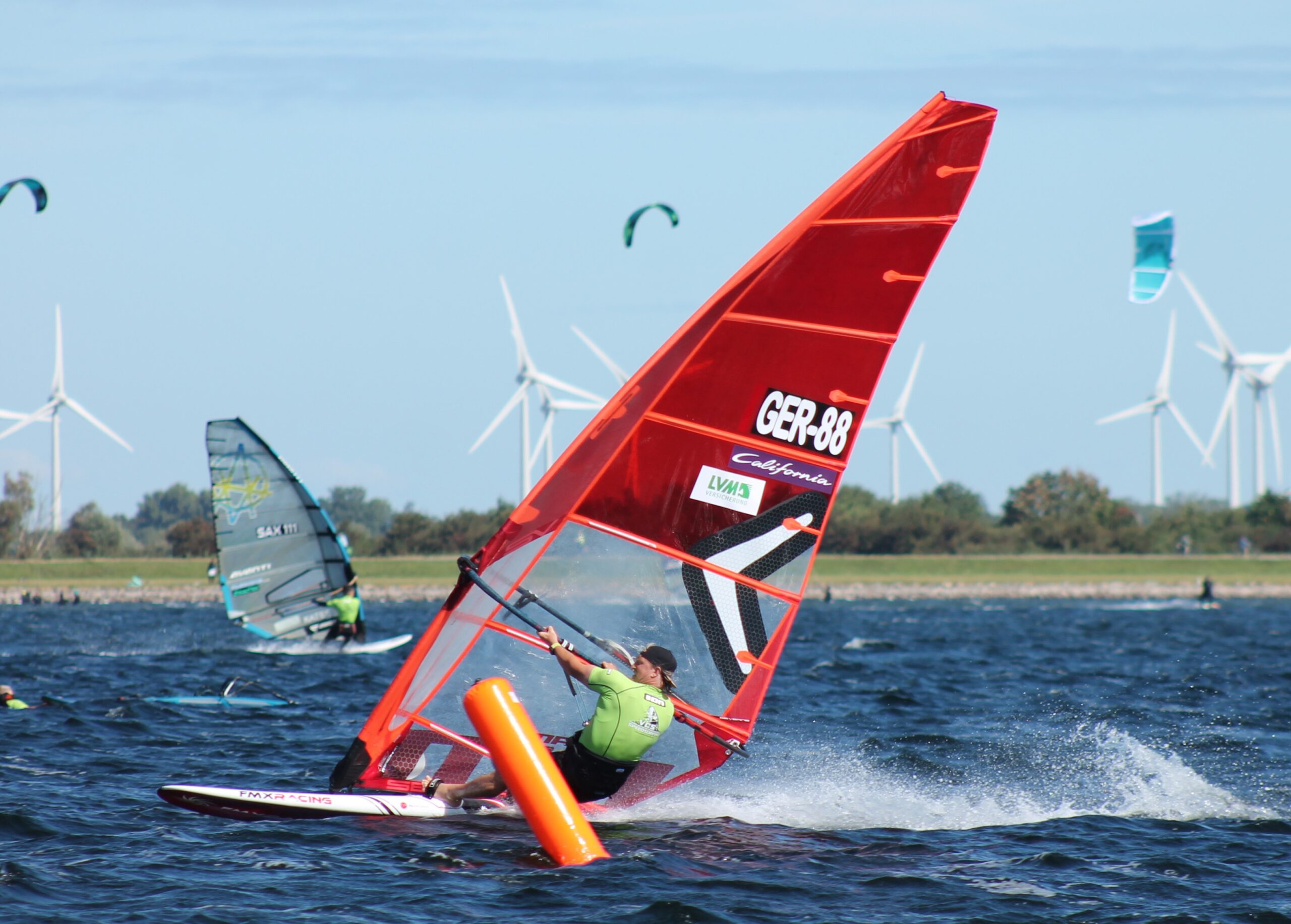 Ein Speedwindsurfer rast über die ostsee. Das Segel ist rot. Im Hintergrund ist ein zweiter Fahrer zu sehen, am Himmel sind Drachen von Kitesurfenden.
