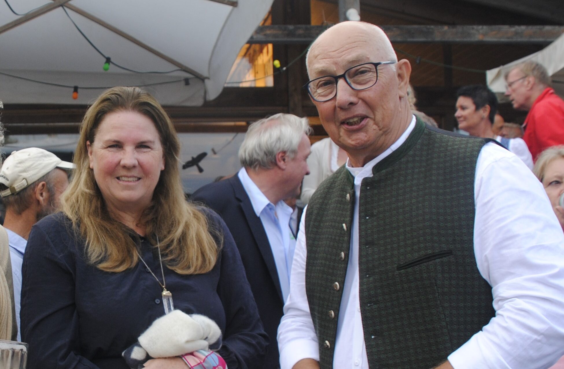 Eine Frau mit offenen Haaren und einem blauen Pullover neben einem älteren Mann mit Brille und Glatze, er trägt eine Weste und ein weißes Hemd ohne Kragen.