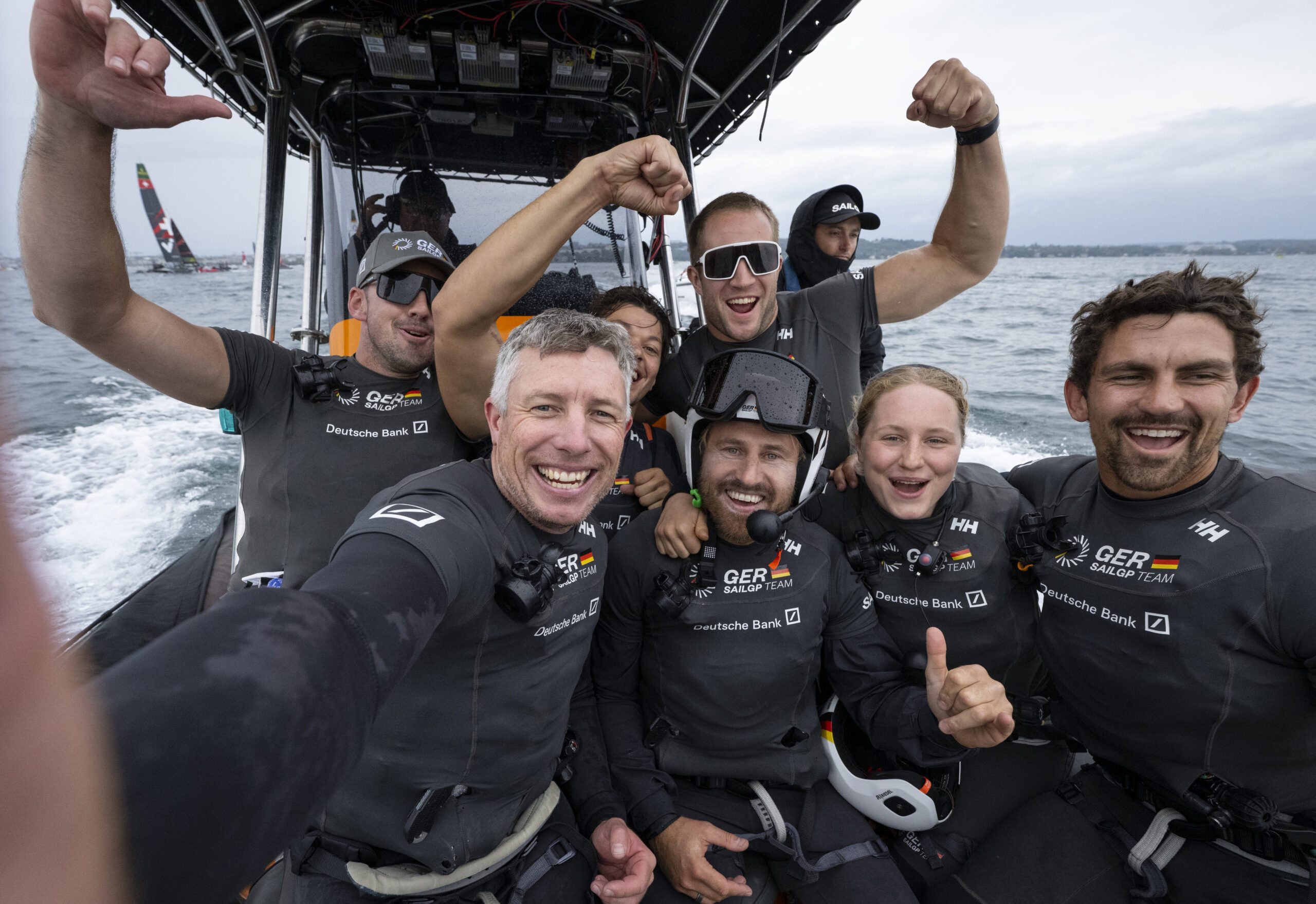 Jubelnde Menschen mit schwarzer Wassersportbekleidung auf einem Motorboot, der Mann in der Mitte trägt einen Helm.