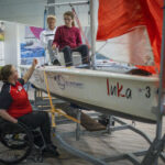 Zwei Segeknde sitzen in einem inklusiven Segelboot, das auf einem Messestand aufgestellt ist. Eine Seglerin im Rollstuhl sitzend erklärt die Funktionen des Bootes.