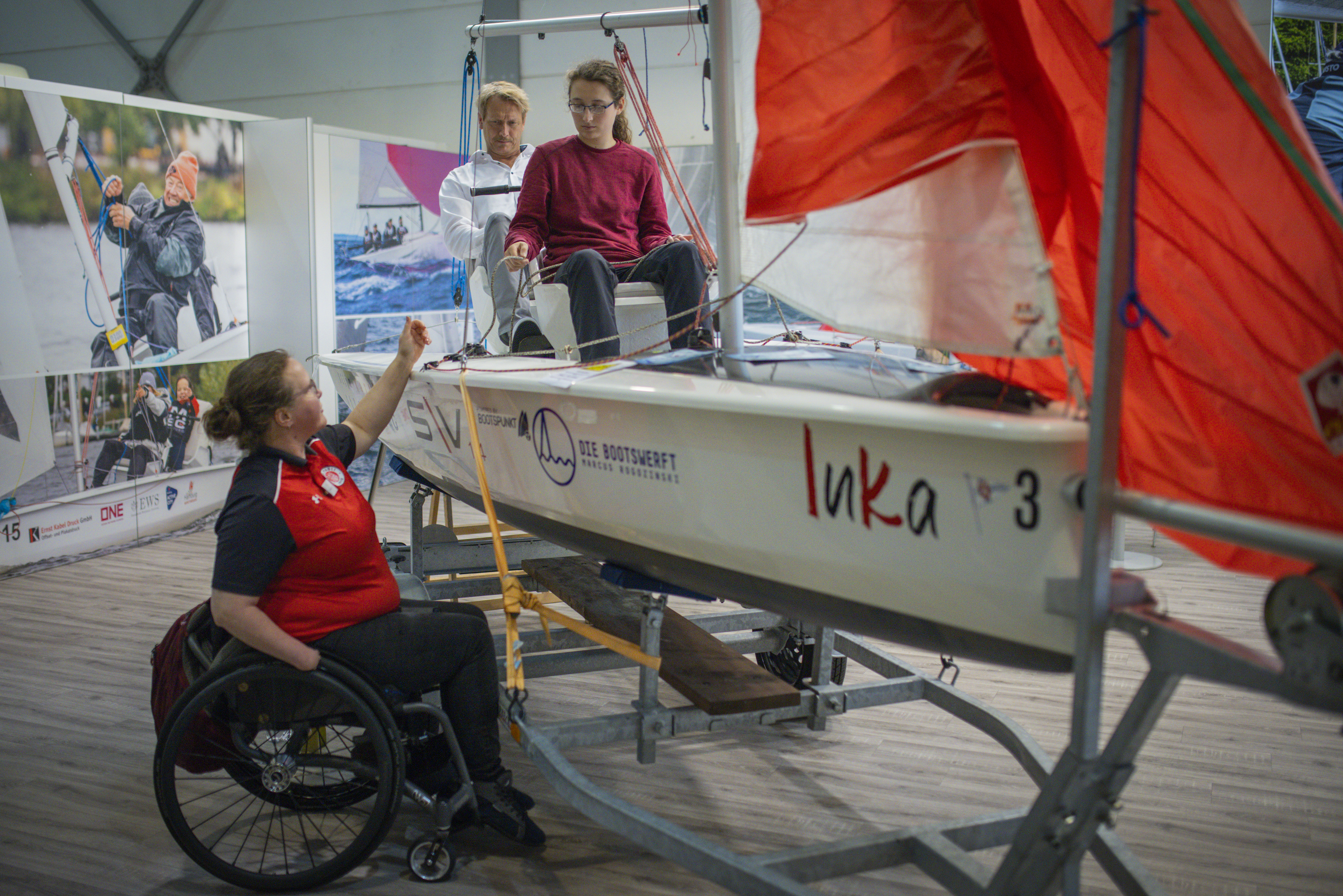 Zwei Segeknde sitzen in einem inklusiven Segelboot, das auf einem Messestand aufgestellt ist. Eine Seglerin im Rollstuhl sitzend erklärt die Funktionen des Bootes.