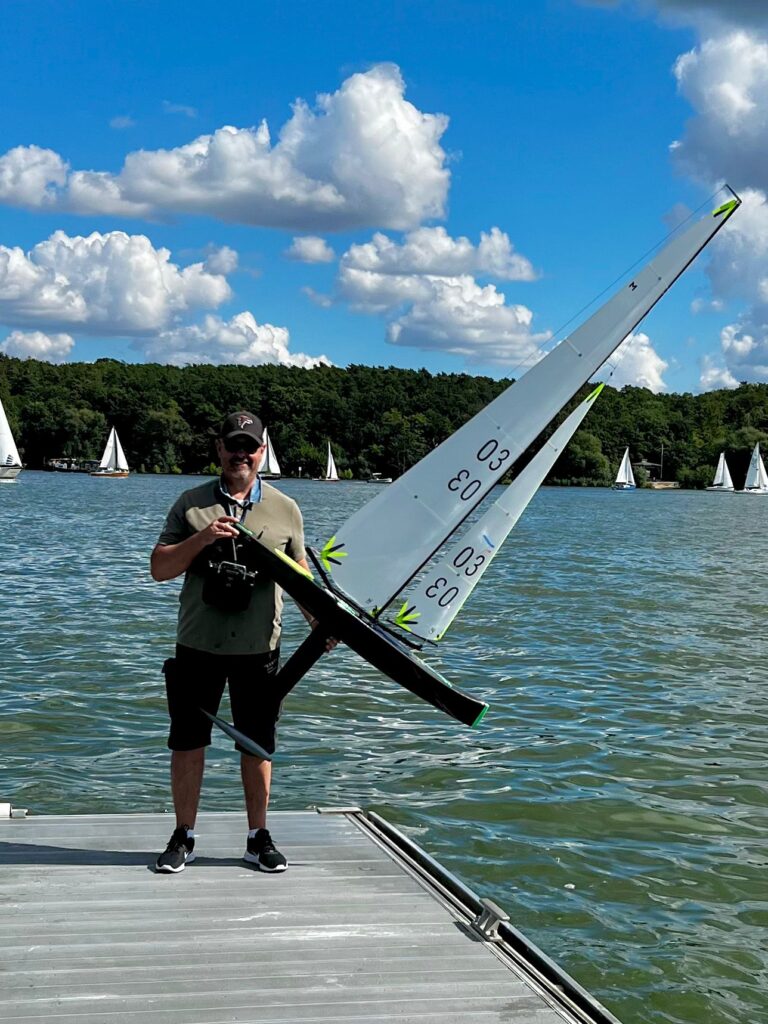 Ein Mann steht am Ende eines Stegs. Im Hintergrund ist Wasser zu sehen. In der Hand hält er eine Modell-Segelyacht. Der Rumpf des Bootes ist zirka 1,20 Meter lang, das Rigg mit Mast und Segeln zirka zwei Meter hoch.
