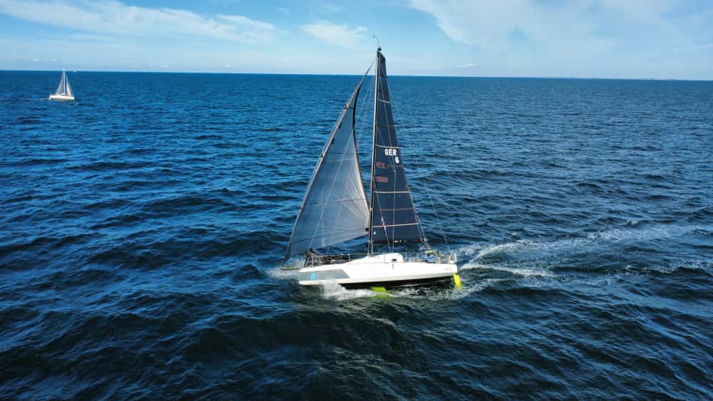 Eine Segelyacht mit einem hellen Vorsegel und einem schwarzen Hauptsegel mit einer Zahl darin, der Rumpf ist weiß, Ruder und Schwert sind neongelb und durch das Wasser zu erkennen.