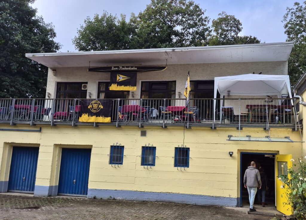 Das Foto zeigt ein gelbes Haus mit Eingang in der unteren Etage. Darüber ist eine Terrasse mit Tischen und Stühlen zu sehen. Am Geländer hängt ein Plakat: 100 Jahre Kanu Club Speyer.