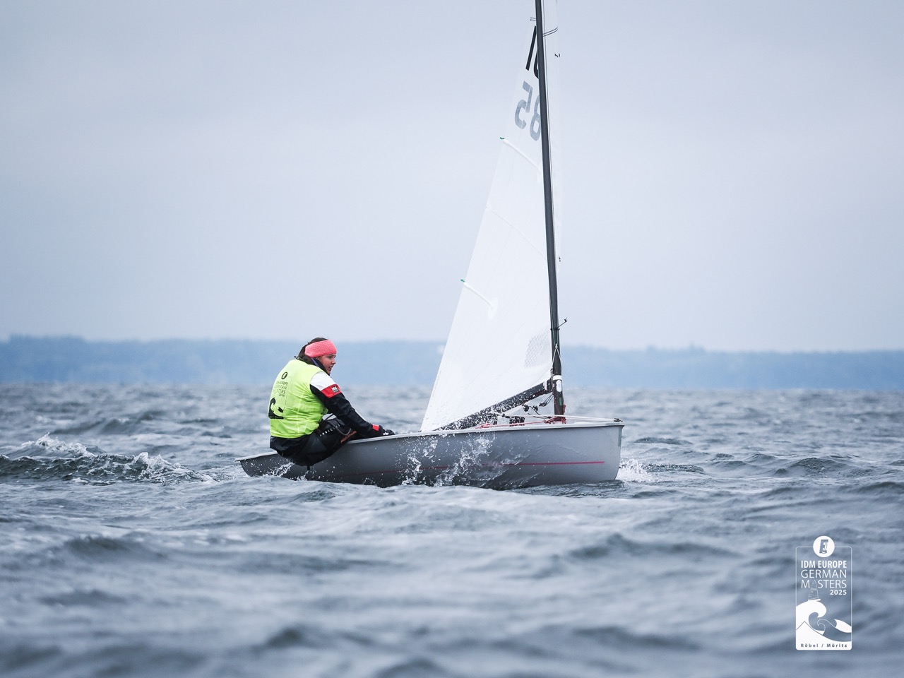 Eine Frau in einem gelben Trikot segelt auf einer Jolle mit einem weißen Segel und einem schwarzen Mast.