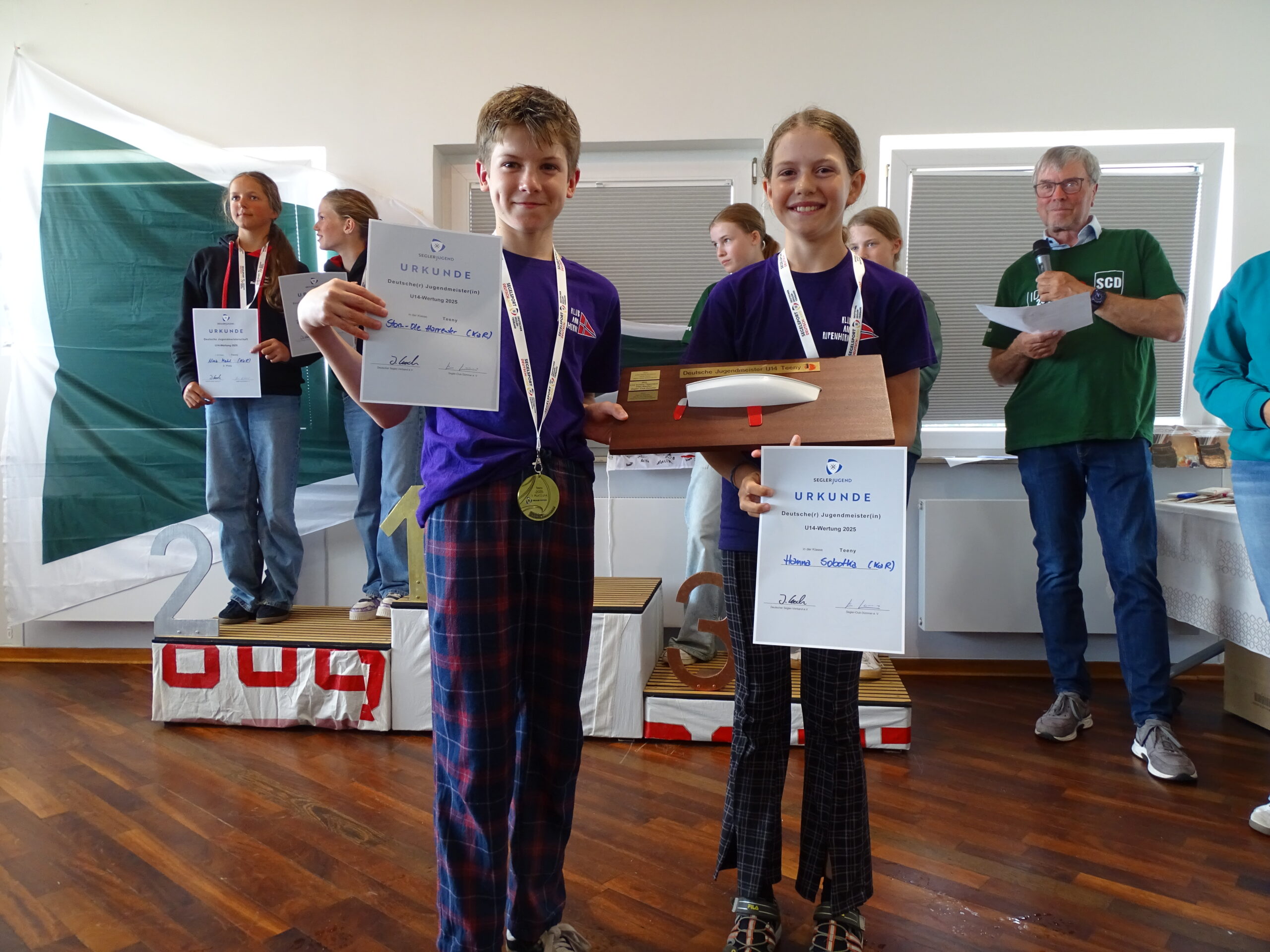 Zwei Jugendliche halten stolz ihre Siegerurkunden in die Kamera, der Junge hat ein blaues T-Shirt an und eine Medaille um den Hals, das Mädchen trägt zusätzlich einen Wanderpreis mit einem Schiff darauf im Arm.