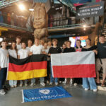14 deutsche und polnische Jugendliche stehen in einem großen Surfshop. Sie lächeln in die Kamera und halten eine deutsche und eine polnische Flagge in der Hand.