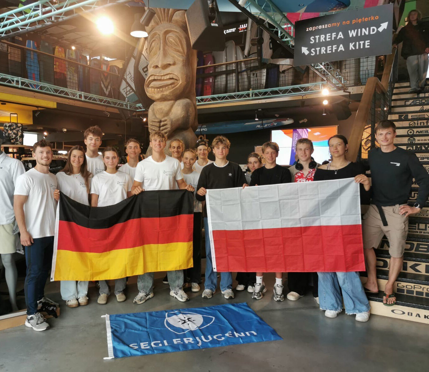 14 deutsche und polnische Jugendliche stehen in einem großen Surfshop. Sie lächeln in die Kamera und halten eine deutsche und eine polnische Flagge in der Hand.