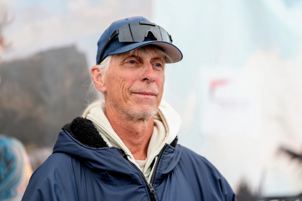Detlef Groebert schaut rechts am Fotografen vorbei und lächelt leicht. Der Hintergrund ist unscharf. Er trägt ein Cap und eine Sonnenbrille auf dem Cap.