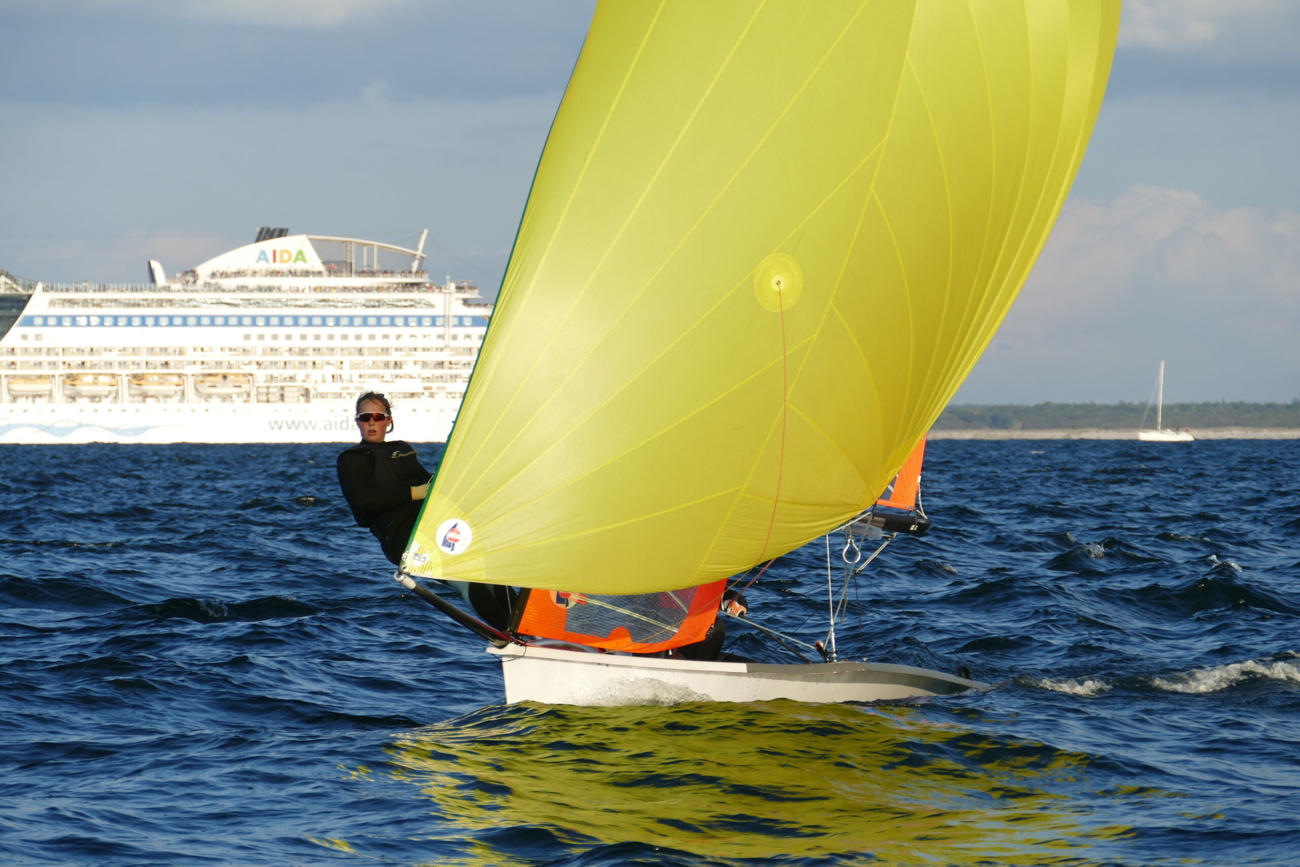 Eine Skiffjolle mit einem gelben Spinnaker vor einem großen Kreuzfahrtschiff.