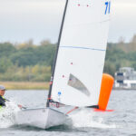Ein Mann mit einem gelben Trikot segelt auf einer Jolle mit einem weißen Segel, neben ihm ist eine orangefarbene Bahnmarke auf dem Wasser.