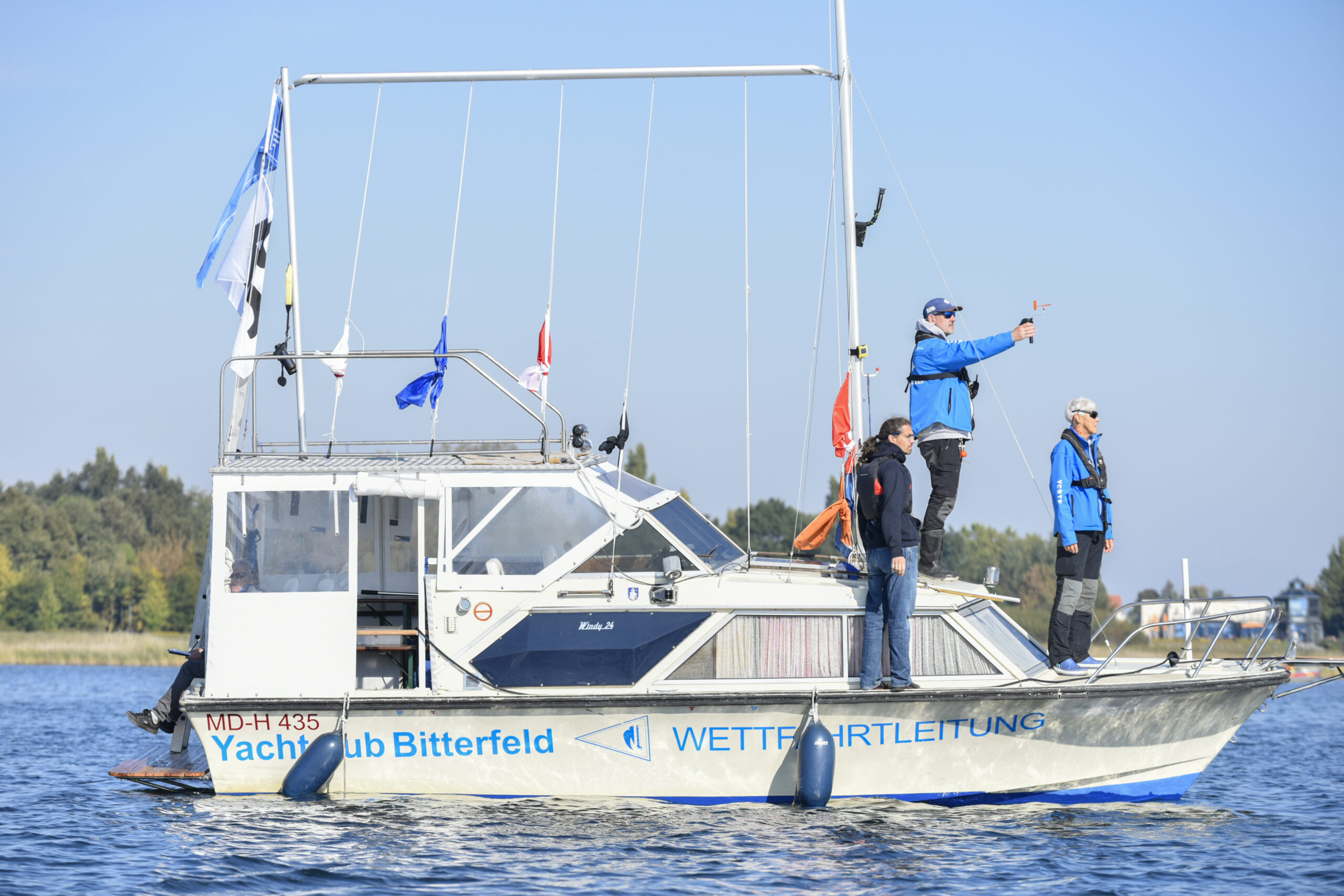 Ein Motorboot mit einem großen Aufbau und vielen Flaggen, die nicht hochgezogen sind. Es wird für die Organisatoren einer Regatta benutzt. Vorne stehen drei Männer mit blauen Jacken, der mittlere misst den Wind.