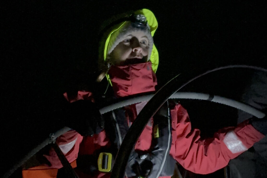 Eine Frau steht am Steuerrad einer Yacht. Es ist Nacht, ihr Gesicht wird von den Bordinstrumenten erhellt. Sie trägt dickes Ölzeug und eine Mütze.