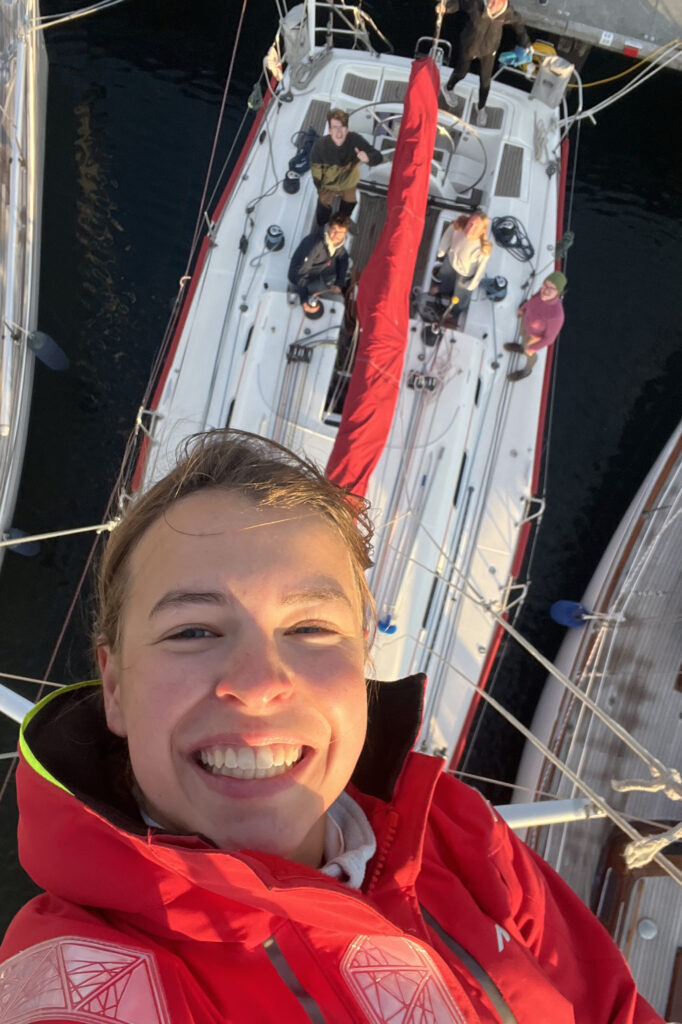 Ein Selfie: Im Vordergrund eine lächelnde Frau, die hoch im Mast einer Segelyacht ist. Tief unter ihr das Boot und die Crew