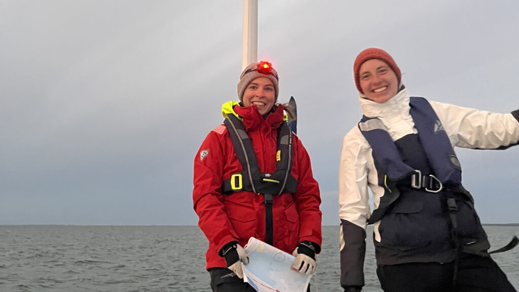 Zwei junge Frauen stehen im Heckkorb einer Yacht und lächeln fröhlich in die Kamera. Eine Frau trägt eine rot leuchtende Stirnlampe.