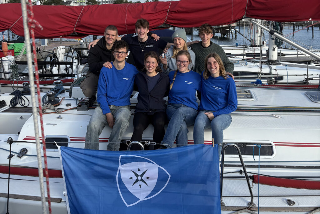 Die DSV-Jugendcrew des Jahrgangs 2025 sitzt auf dem Aufbau der Yacht und lächelt in die Kamera.