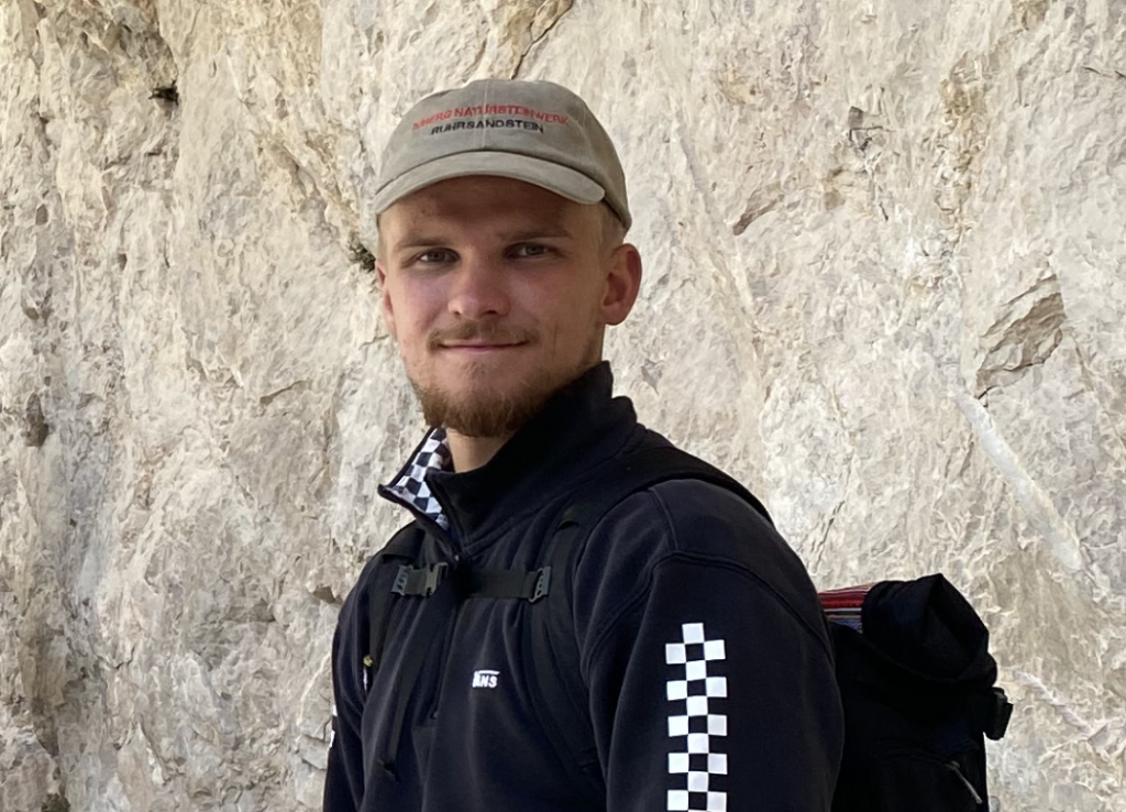 Konrad Struwe steht vor einer Felswand und schaut in die Kamera. Er trägt ein Cap und einen Rucksack