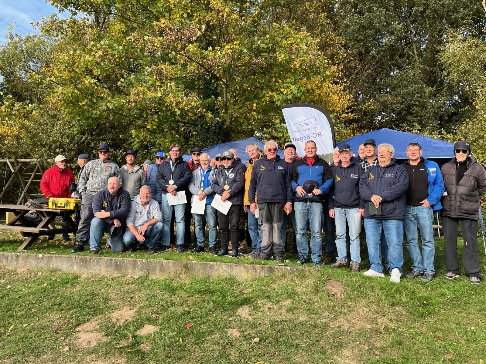 Viele Männer die auf einer grünen Wiese stehen, sie nehmen gemeinsam an einer Regatta für ferngesteuerte Yachten teil.