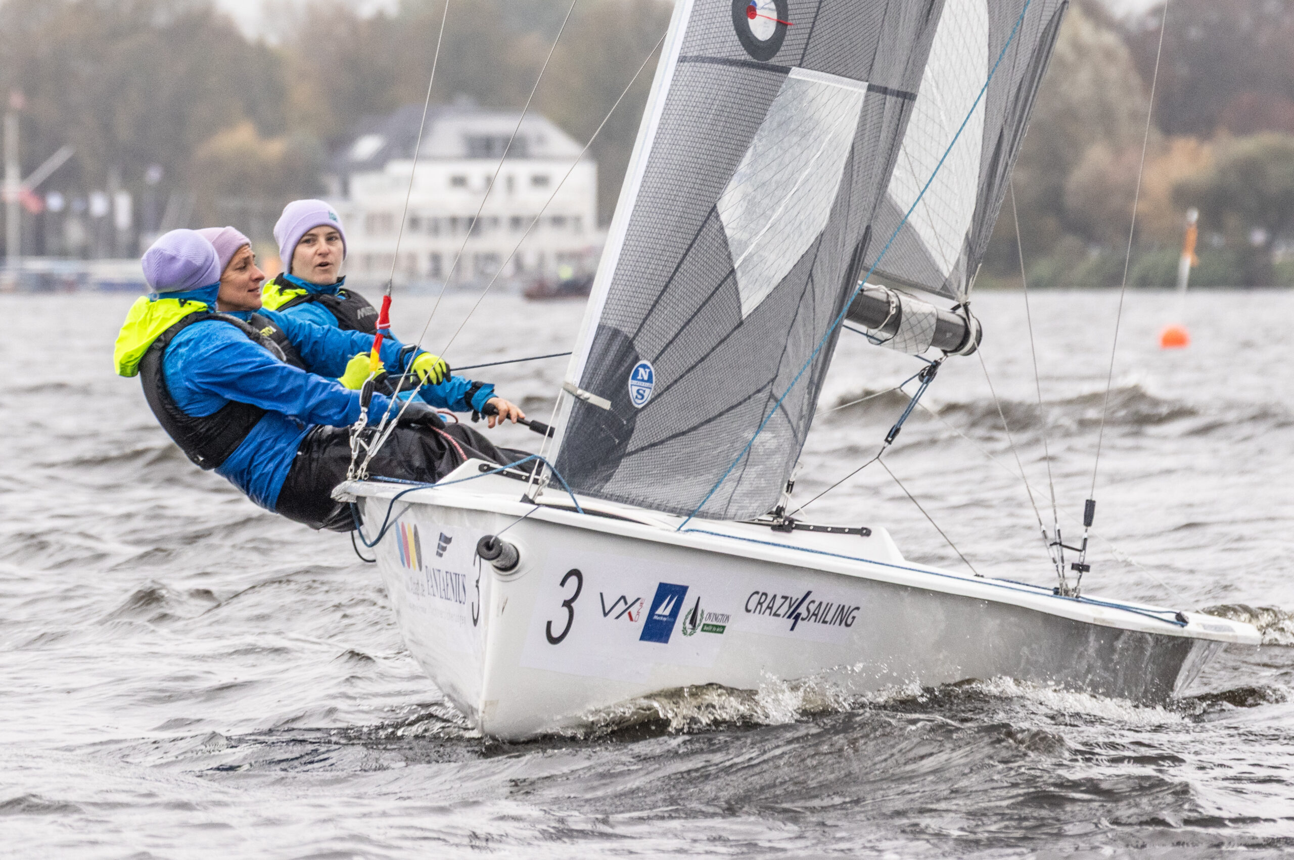 Drei Frauen segeln auf einer weißen Kielschwertyacht, sie tragen blaues Ölzeug. Das Wetter ist schlecht, im Hintergrund ist das Gebäude des Norddeutschen Regatta Verein an der Alster zu erkennen.