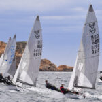 Mehrere Gleitjollen vom Typ 505er auf einer Regatta, links im Bild eine rote Bahnmarkierung, im HIntergrund Felsen.