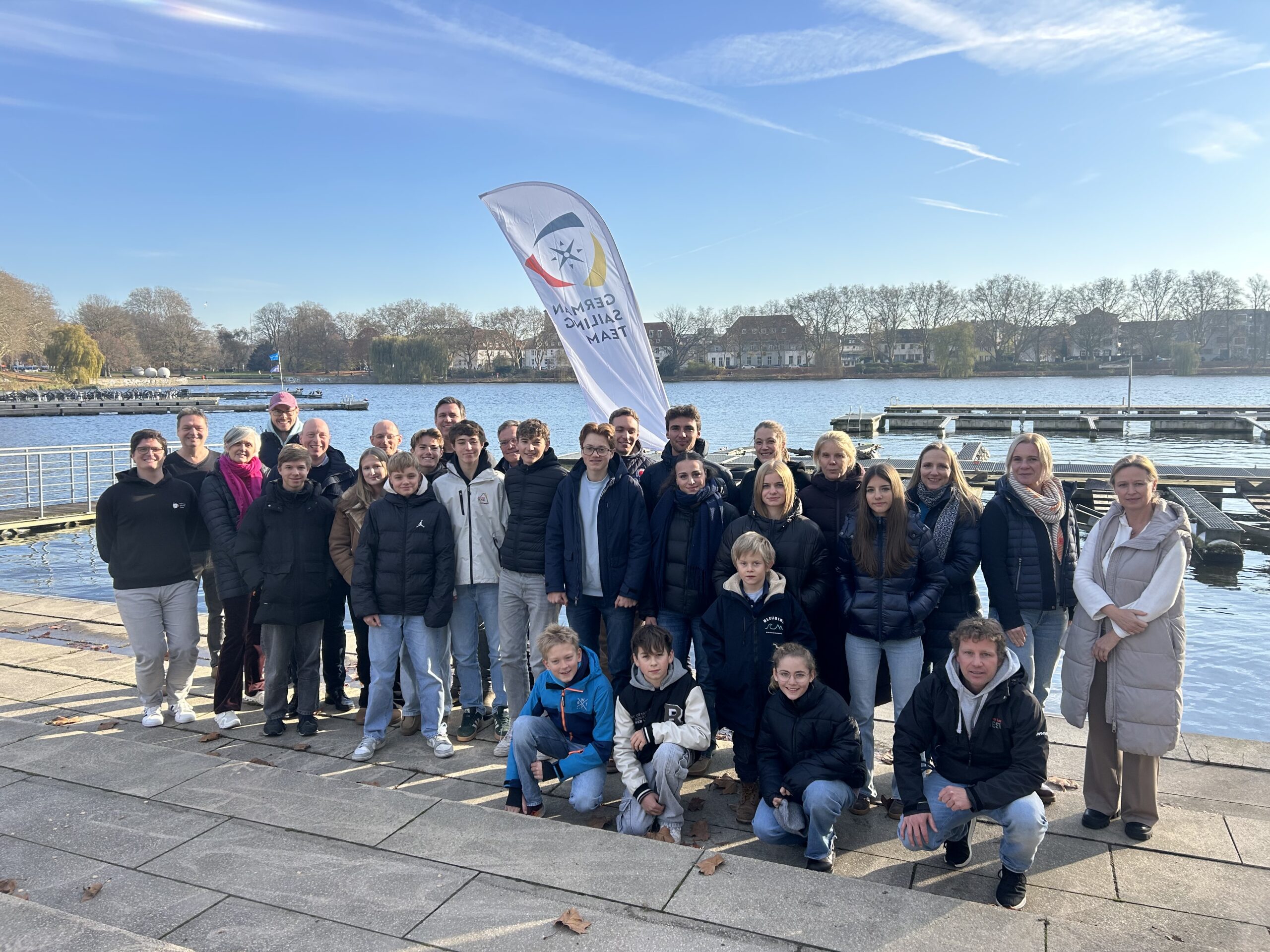 Mehrere Jugendliche, Eltern und Betreuer stehen vor einem See im Winter bei Sonnenschein, dahinter ist die Flagge des Deutschen Segler-Verbandes zu erkennen.