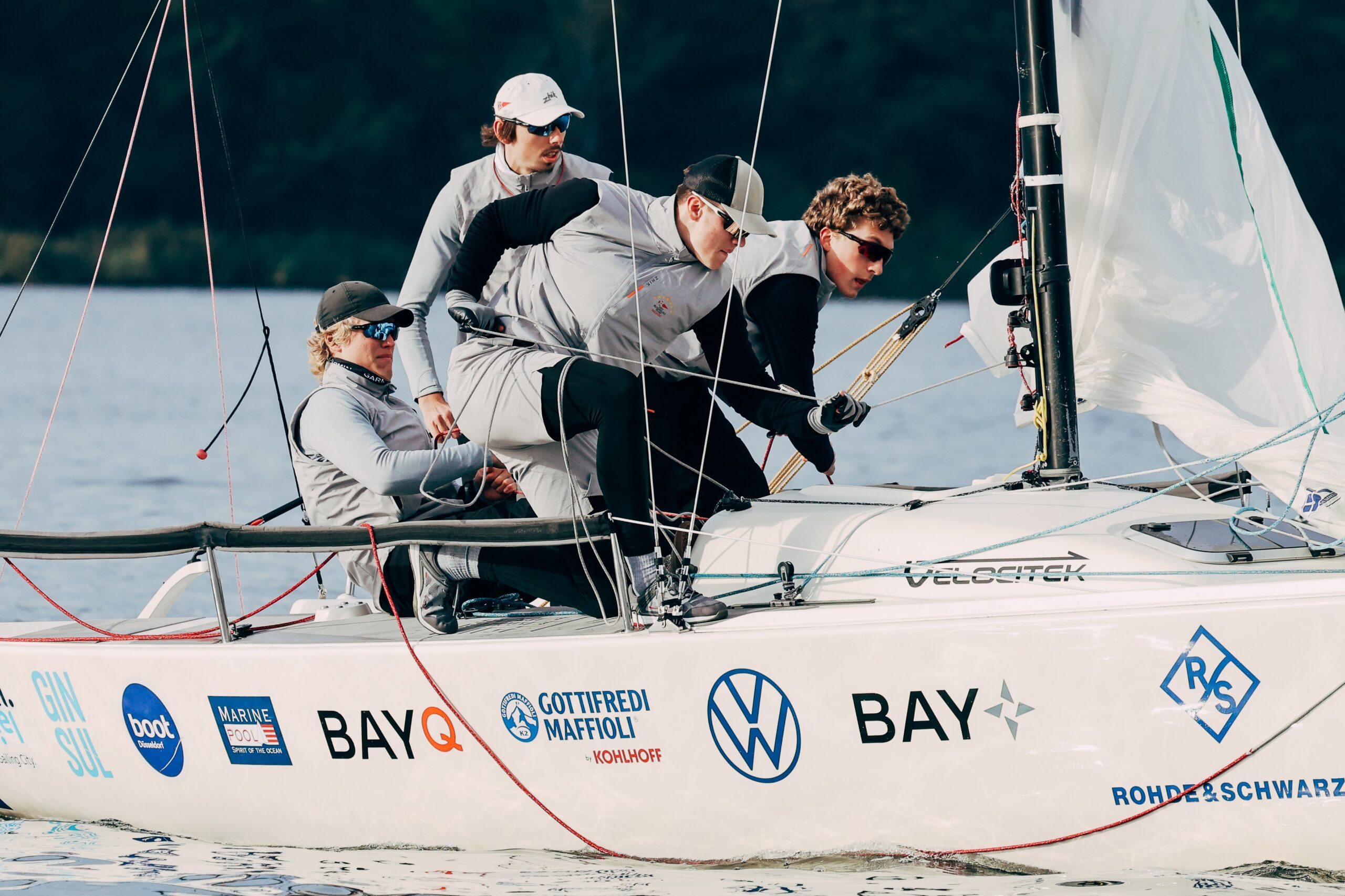 Vier Männer mit grauen Westen und Caps auf einem sportlichen Segelboot mit weißem Rumpf, das mit den Namen zahlreicher Sponsoren beklebt ist.
