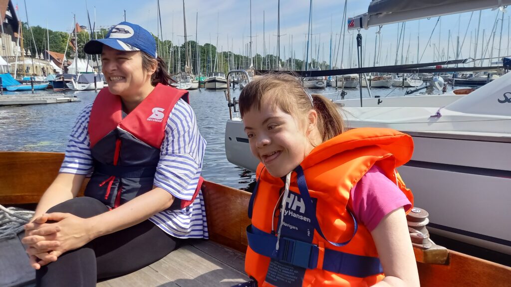 Ein junges Mädchen mit geistigem Handicap freut sich an Bord eines Segelbootes. Sie wird von einer vertrauten Betreuerin begleitet.