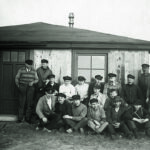 Historisches Bild: Viele Männer mit Hut und Pfeife stehen und sitzen vor einem kleinen Holzhaus