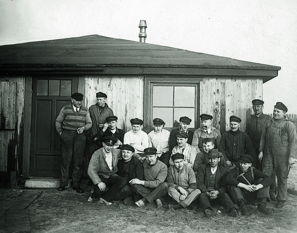 Historisches Bild: Viele Männer mit Hut und Pfeife stehen und sitzen vor einem kleinen Holzhaus