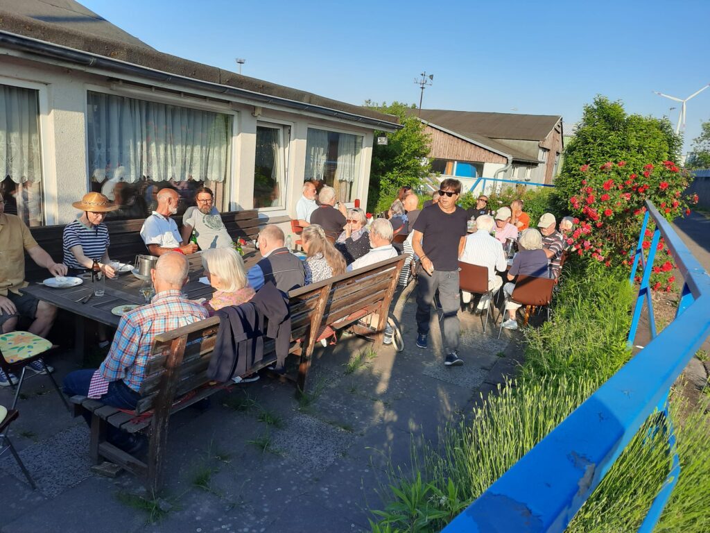 Menschen sitzen gesellig vor dem Clubhaus des SCOe zusammen, die Sonne scheint