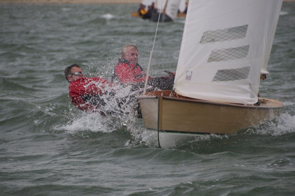 zwei Männer segeln ein Boot vom Typ Sharpie bei viel Wind und müssen sich stark raushängen, um das Boot zu stabilisieren