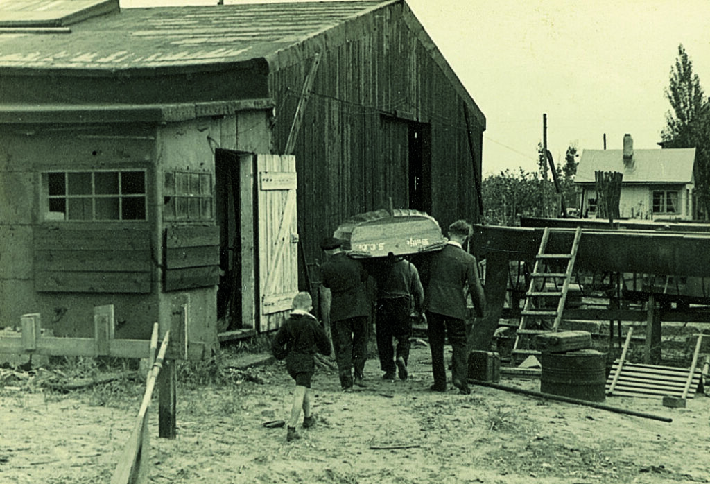 Historisches Bild: Ein Bootsschuppen des SCOe. Drei Männer tragen eine Jolle hinein, ein Junge geht hinterher