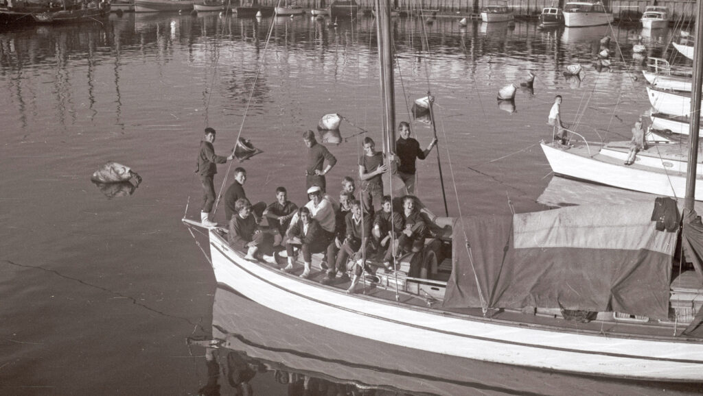 Ein historisches schwarzweiß Foto zeigt eine altes Boot mit Jugendlichen darauf.