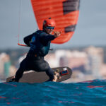 Der Kitesurfer Jan Vöster zeigt auf seinem Board stehend Hang Loose in die Kamera